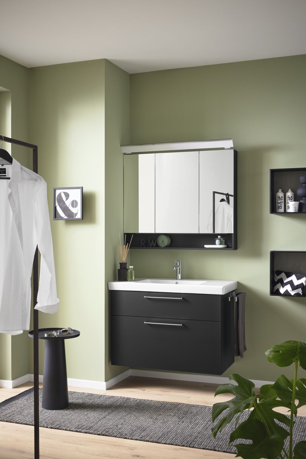 Badkamer met lente groene muur en zwarte meubels, witte wasbak en spiegelkastje met verlichting van boven.